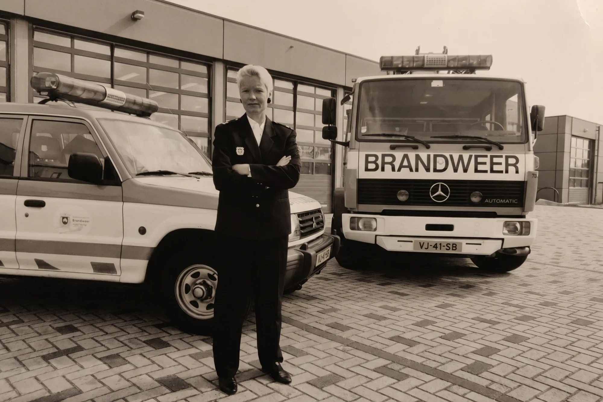 Het interieur van een Nederlandse brandweerkazerne &mdash; een rode brandweerwagen geparkeerd in een schone garage, brandweerjassen aan de linkerwand, daglicht door grote ramen.
